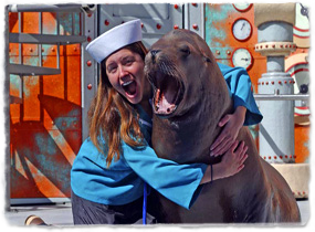 A trainer wearing a sailor cap hugs a sea lion while laughing.