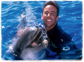 A trainer pets a dolphin while the two pose in the water.